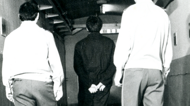 Ein Häftling wird von zwei Aufsehern einen Gang entlang geführt.