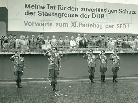 Vor dem Podium sind fünf Soldaten in Uniform zum militärischen Gruß aufgestellt. Vor dem zweiten Soldaten von links steht ein Mikrofon. Auf der Tribüne befinden sich mehrere Männer in ziviler Kleidung und in Uniform sowie eine Frau. Über den Personen auf der Tribüne steht "Meine Tat für den zuverlässigen Schutz der Staatsgrenze der DDR! Vorwärts zum XI. Parteitag der SED!".