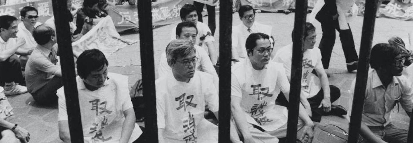 Auf dem Schwarz-Weiß-Bild zu sehen sind männliche auf dem Boden sitzende Demonstranten in heller Kleidung.
