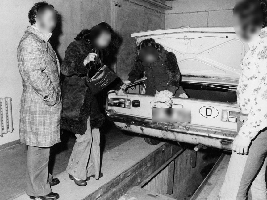 Das Bild zeigt ein Westberliner Fahrzeug, das Stasi-Mitarbeiter von hinten fotografierten. Das Fahrzeug steht ebenerdig auf einer Art Hebebühne. Der Kofferraum ist geöffnet. Ein Mann und eine Frau stehen links vom Fahrzeug. Rechts vom PKW ist der vermeintliche Fahrer zu erkennen. Eine weitere Person steigt gerade aus dem Kofferraum.