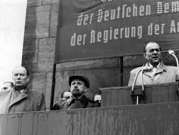 Das Bild zeigt Wilhelm Zaisser, Walter Ulbricht, Otto Grotewohl während einer Kundgebung auf einer Bühne. Grotewohl spricht gerade in ein Mikrofon.