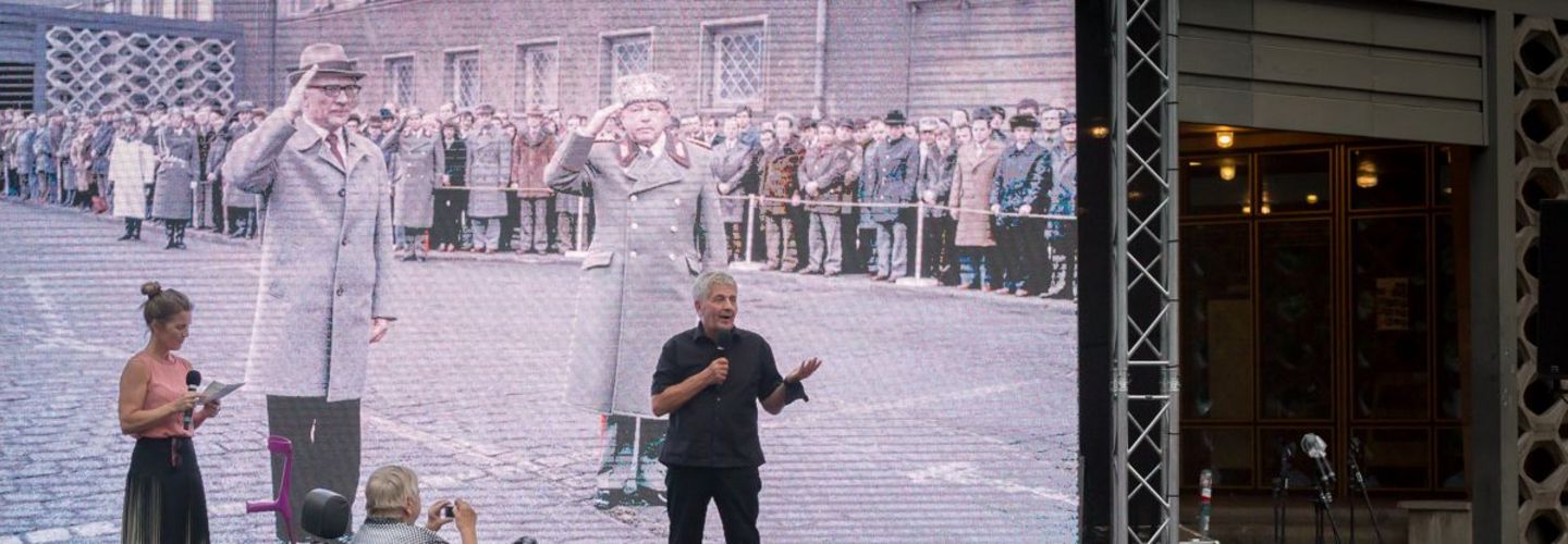 Im Hintergrund spricht Roland Jahn zu einem im Vordergrund befindlichen Publikum. Er steht vor einem großen Screen im Hintergrund, auf dem Erich Honecker und Erich Mielke vor Haus 1 zu sehen sind.