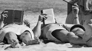 Zwei Personen liegen am Strand und lesen jeweils ein Buch. Im Hintergrund ist das Meer zu erkennen und eine Person hält dazu eine Zigarette in der Hand