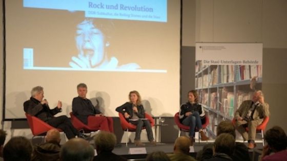 Auf einer Bühne sitzen vier Personen in roten Sesseln. Im Hintergrund ist auf einer Leinwand das Cover des Dokumentenhefts 'Rock und Revolution' zu sehen.