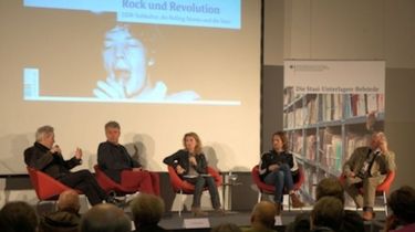 Auf einer Bühne sitzen vier Personen in roten Sesseln. Im Hintergrund ist auf einer Leinwand das Cover des Dokumentenhefts 'Rock und Revolution' zu sehen.