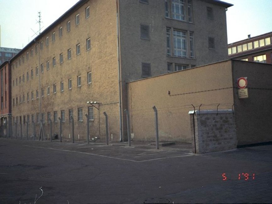 Im Hintergrund ist die mit Stacheldrahtzäunen umgebene Haftanstalt zu sehen. Davor befindet sich eine Straße.
