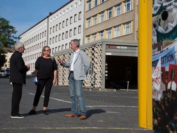 Roland Jahn, Katrin Budde und Frank Ebert stehen auf dem zentralen Hof des MfS-Geländes. Im Hintergrund ist der Eingang zu Haus 1 zu erkennen.