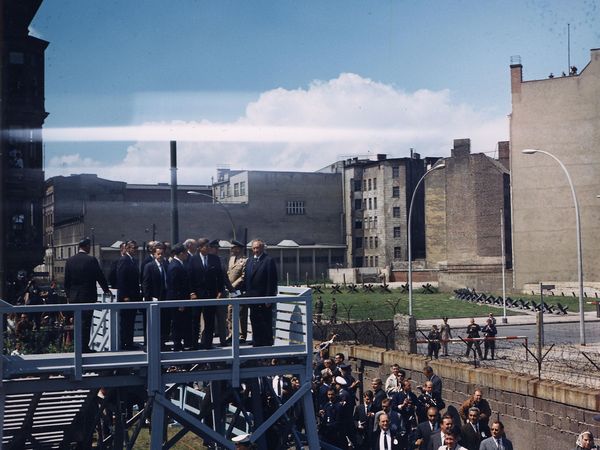 John F. Kennedy steht in einer kleinen Gruppe auf einer Plattform am Checkpoint Charlie unmittelbar an der Berliner Mauer mit Blick auf den Ostteil der Stadt. Neben ihm steht Bundeskanzler Konrad Adenauer. Unten befindet sich eine Menschenansammlung, darunter Journalisten. 