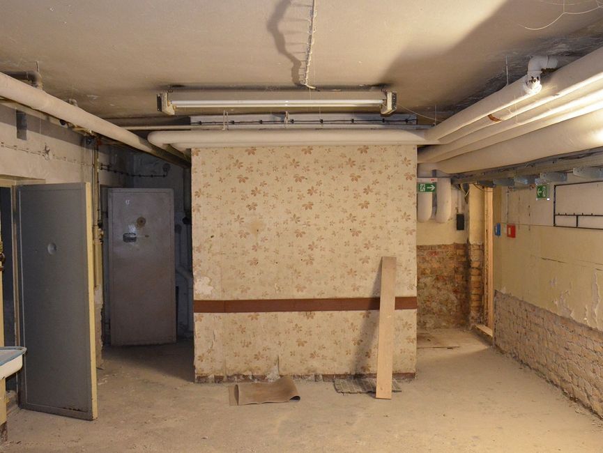 Ein Kellergewölbe. Links sind zwei Zellentüren geöffnet, rechts ist ein Ausang. Unter der Decke laufen Rohre entlang.