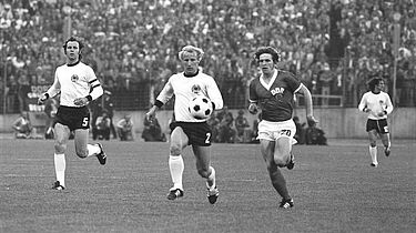 Fußballszene, bei der der DDR-Stürmer Martin Hoffmann und der Verteidiger der westdeutschen Nationalmannschaft Berti Vogts dem Ball nachstürmen. Links ist Franz Beckenbauer, im Hintergrund Wolfgang Overath zu sehen (beide Bundesrepublik).