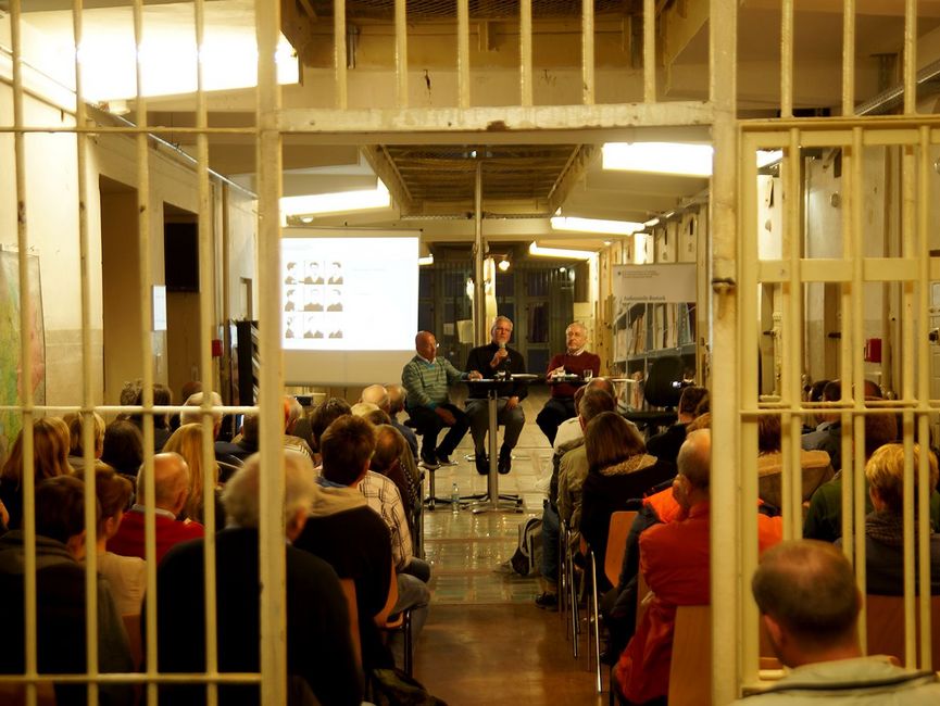 Im Hintergrund sind drei Vortragende zu erkennen, die vor einer kleinen Leinwand sitzen. Vor ihnen sitzt ein Publikum. Den Vordergrund bilden Gefängnisgitter.
