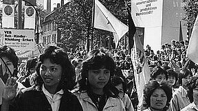 Ein Demonstrationszug mit vielen vietnamesischen Arbeitskräften auf den Straßen Erfurts. Im Hintergrund ist ein Häuserzug und eine Tribüne zu sehen. Etliche Fahnen der DDR werden geschwenkt und das Schild des Volkseigenen Betriebs (VEB) Reh-Kinder-Kleidung-Erfurt ist im Demonstrationszug zu erkennen.