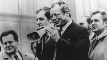 Zu sehen ist eine Gruppe von sechs Männern. Im Vordergrund steht Willy Brandt mit einem Megafon in der Hand, in das er gerade hineinspricht. Am linken Bildrand sieht man Günter Guillaume mit Brille und gemusterter Krawatte.