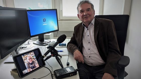 Das Bild zeigt Günter Bormann an seinem Arbeitsplatz. Vor ihm befindet sich ein Mikrofon und ein Tablet, mit welchem Bormann gerade ein Videotelefonat mit dem Journalisten Maximilian Schönherr führt.