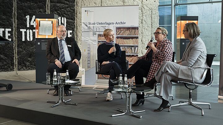 Drei Frauen und ein Mann sitzen auf einer Bühne und sprechen miteinander. Im Hintergrund sind eine Obejktstele und ein Banner mit der Aufschrift 'Stasi-Unterlagen-Archiv' zu sehen.