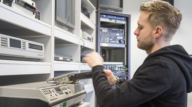 Ein Archivmitarbeiter steht vor einem Regal mit mehreren Videorekordern und -abspielgeräten. Er steckt gerade eine Videokassette in den Schlitz eines Videogerätes.