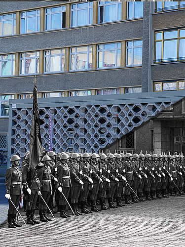 Das Bild ist schräg in der Mitte geteilt: Die Hälfte oben links zeigt ein aktuelles Foto der Vorderfront von Haus 1 mit dem Sichtschutz. Die Hälfte unten rechts zeigt eine Reihe von Soldaten in Uniformen, vor denen Erich Mielke steht.