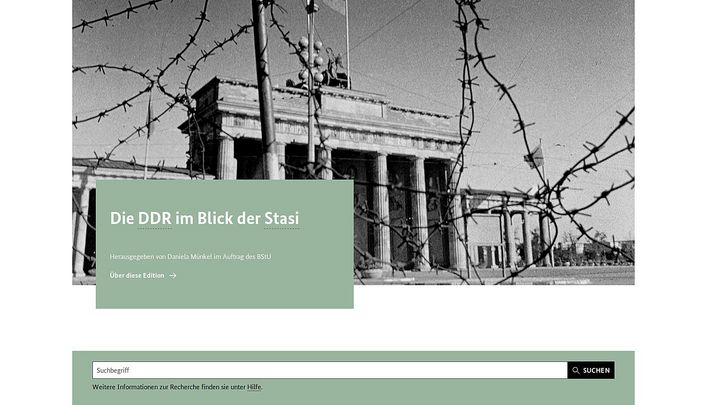 Das Bild zeigt einen Ausschnitt der Startseite der ZAIG-Datenbank 'DDR im Blick der Stasi'. Zu sehen ist ein Schwarz-Weiß-Bild des Brandenburger Tors, das etwas durch Stacheldraht im Vordergrund verdeckt wird. Darüber befindet sich ein mintgrün hinterlegtes Textfeld mit dem Text: ' Die DDR im Blick der Stasi Herausgegeben von Daniela Münkel im Auftrag des BStU'