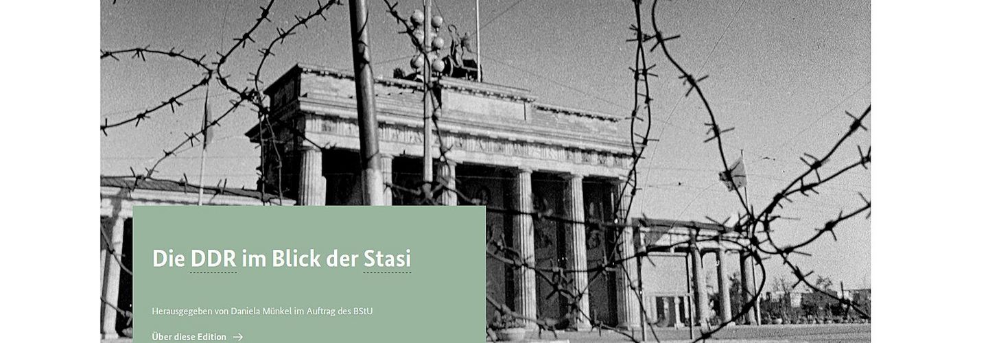 Das Bild zeigt einen Ausschnitt der Startseite der ZAIG-Datenbank 'DDR im Blick der Stasi'. Zu sehen ist ein Schwarz-Weiß-Bild des Brandenburger Tors, das etwas durch Stacheldraht im Vordergrund verdeckt wird. Darüber befindet sich ein mintgrün hinterlegtes Textfeld mit dem Text: ' Die DDR im Blick der Stasi Herausgegeben von Daniela Münkel im Auftrag des BStU'