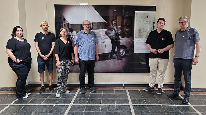 Sechs Personen stehen vor einer Wand, auf der ein großformatiges Foto von Wolf Biermann und vom Aktendeckel des Zentralen Operativen Vorgangs 'Lyriker' zu sehen ist.
