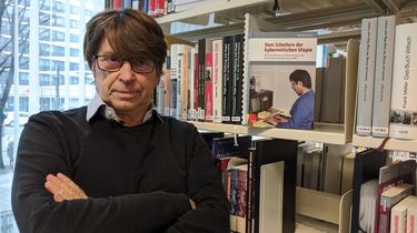 Dr. Christian Booß steht neben einem Bücherregal, in dem sein Buch Dr. Christian Booß neben seiner Publikation 'Das Scheitern der kybernetischen Utopie' zu erkennen ist.