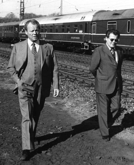 Das schwarz-weiße Lichtbild zeigt Bundeskanzler Willy Brandt, der neben Bahngleisen entlang geht. Schräg hinter ihm läuft Günter Guillaume mit Sonnenbrille. Beide tragen Anzug. Im Hitnergrund steht ein Zug.] 