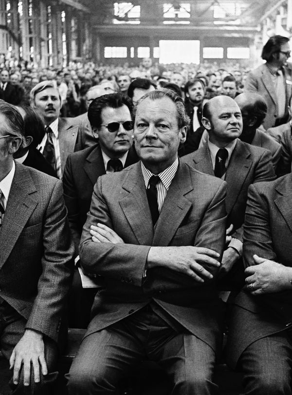 Aufnahme mehrerer Sitzreihen bei einer Wahlkampfveranstaltung. Vorn sitzt Bundeskanzler Willy Brandt mit verschränkten Armen. Schräg hinter ihm sitzt Günter Guillaume. Er trägt eine schwarze Sonnenbrille.