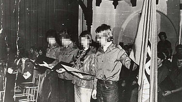 Das Bild zeigt vier Jugendliche auf einer Bühne stehend. Ganz rechts außen befindet sich eine Fahne der FDJ.
