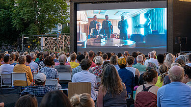 Menschen sitzen vor einer großen Leinwand und sehen den Spielfilm 'Leander Haußmanns Stasikomödie'.
