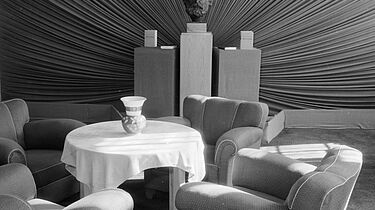 Das Foto zeigt im Vordergrund des Bildes eine Sitzgruppe mit vier Sesseln um einen Tisch. Im Hintergrund ist eine Karl-Marx-Büste aufgestellt. Das Tepetenmuster erweckt den Eindruck, als ob die Büste im Mittelpunkt steht und strahlt.
