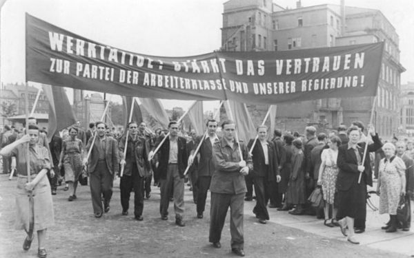 Arbeiterinnen und Arbeiter auf einem Demonstrationszug halten ein Transparent hoch mit der Aufschrift: "Werktätige! Stärkt das Vertrauen zur Partei der Arbeiterklasse und unserer Regierung"