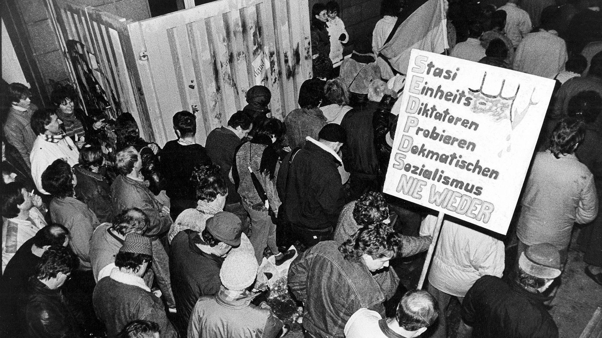 Das Bild zeigt Menschen, die durch ein geöffnetes Tor strömen und Plakate bei sich tragen. Auf einem ist zu lesen: Stasi / Einheits / Diktatoren / Probieren / Demokratischen / Sozialismus / Nie Wieder "