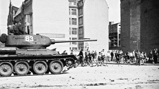 Eine Gruppe von Menschen mit Fahrrädern beobachtet einen sowjetischen Panzer mit zwei Soldaten in offener Luke. Die Szene spielt sich auf einem offenen Platz ab, im Hintergrund sind Wohnhäuser zu sehen.