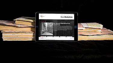 Das Bild zeigt die auf einem Tablet geöffnete Startseite der Stasi-Mediathek. Das Tablet lehnt an einigen Aken.