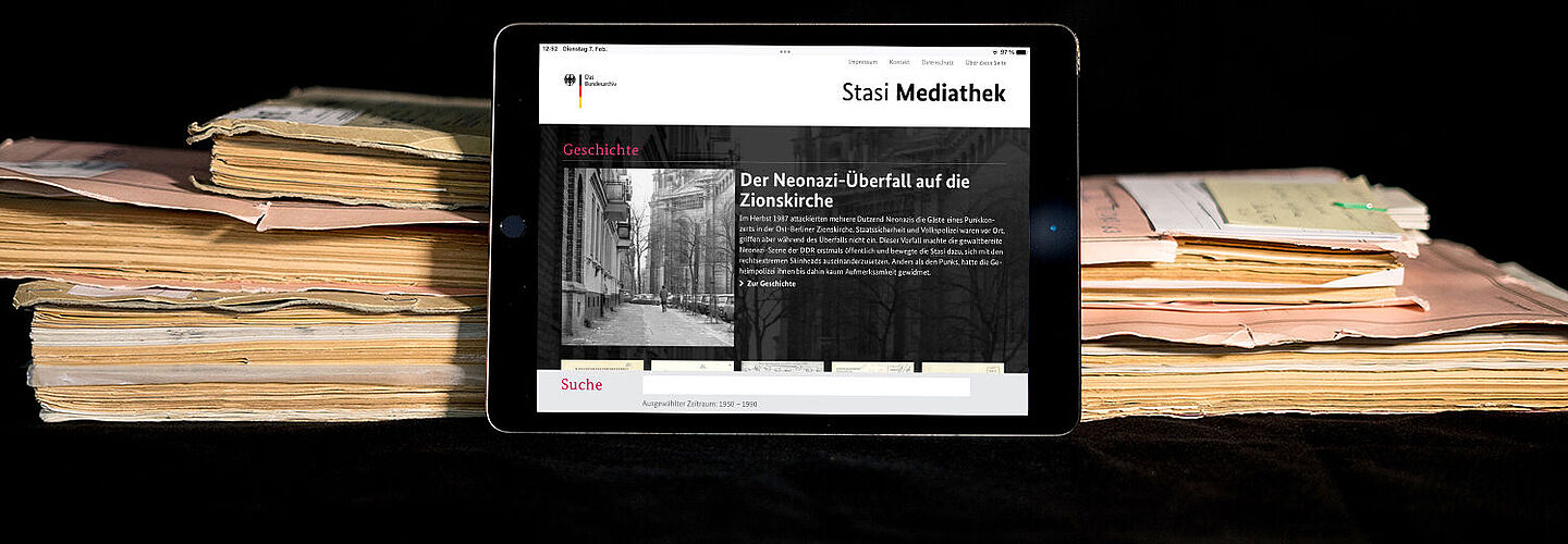 Das Bild zeigt die auf einem Tablet geöffnete Startseite der Stasi-Mediathek. Das Tablet lehnt an einigen Aken.