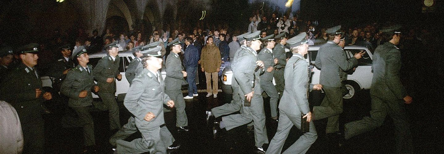 Das Bild zeigt mehrere uniformierte Polizisten, die an stehenden Menschen vorbei eine Straße entlang rennen.