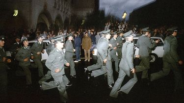 Das Bild zeigt mehrere uniformierte Polizisten, die an stehenden Menschen vorbei eine Straße entlang rennen.