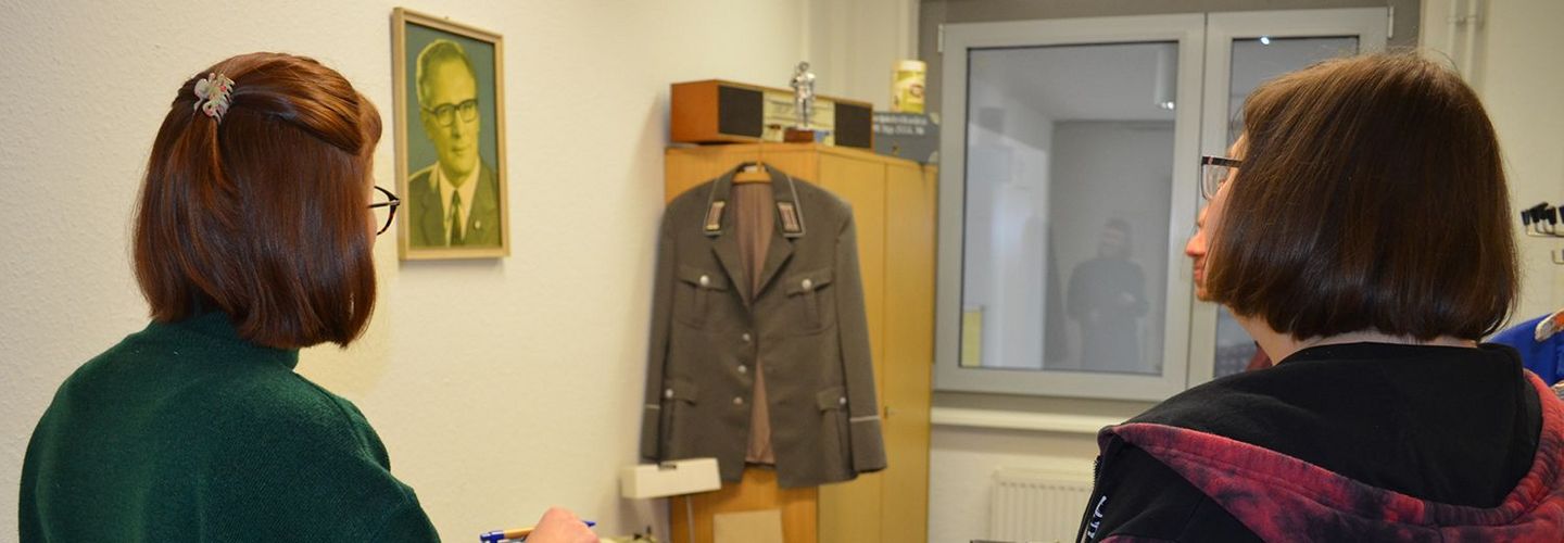 Drei junge Menschen stehen in einem Raum und sehen sich Objekte mit Stasi-Bezug an.