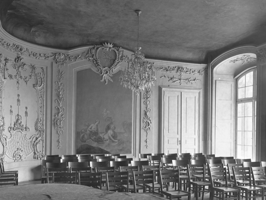 Das schwarz-weiße Lichtbild wurde in dem "Gartensaal" des Schloss Nischwitz, Gemeinde Thalwitz, aufgenommen. Der durch zwei Wandgemälde geschmückte Saal, im Stil des spätbarock mit Stuck versehen, ist bestuhlt. Auf der rechten Wandseite ist ein Bodentiefes Fenster. Es handelt sich um ein schwarz-weißes Lichtbild.