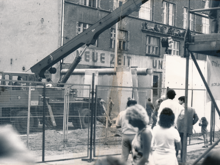 Vor dem Gebäude der "Neuen Zeit" vom Union Verlag, steht ein Schwerlastkran. Des weiteren sind Teile der Berliner Mauer auf dem schwarz-weißen Lichtbild zu sehen. Männer in Uniformen werden durch einen Bauzaun von Schaulustigen getrennt. Das Foto war mittig zerrissen worden und ist nun manuell rekonstruiert.