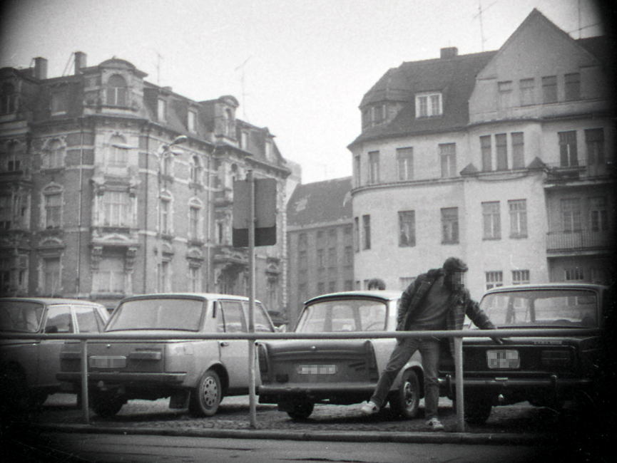 Die Aufnahme zeigt einen öffentlichen Parkplatz vor größeren Mehrfamilienhäusern in einer städtischen Umgebung. Ein Mann hantiert an der Kofferraumhaube eines Pkw.
