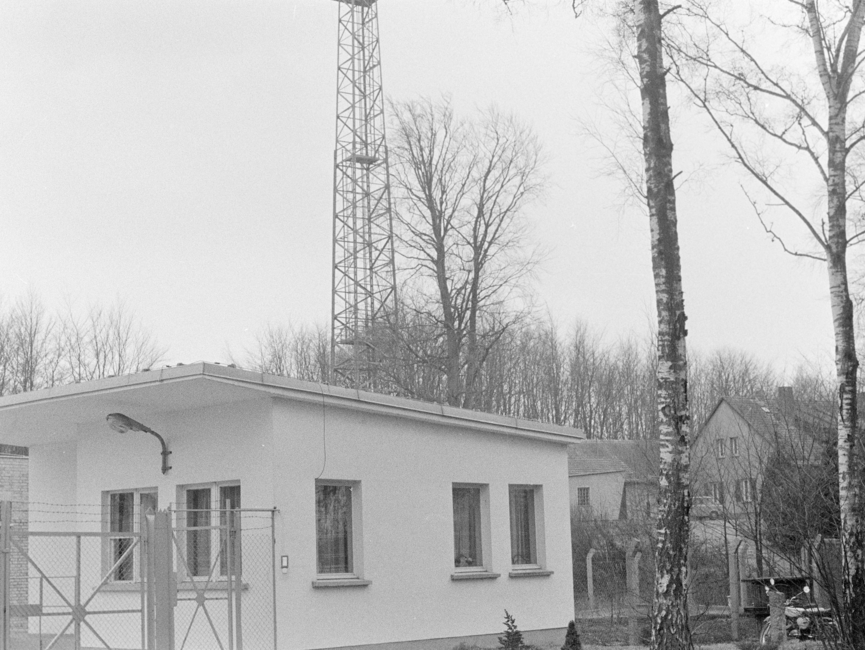 Dieses schwarz-weiße Lichtbild zeigt einen hellen Bungalow auf einem eingezäunten Grundstück, das zusätzlichen mit Stacheldraht geschützt wird. Im Hintergrund ist ein Funkmast erkennbar.