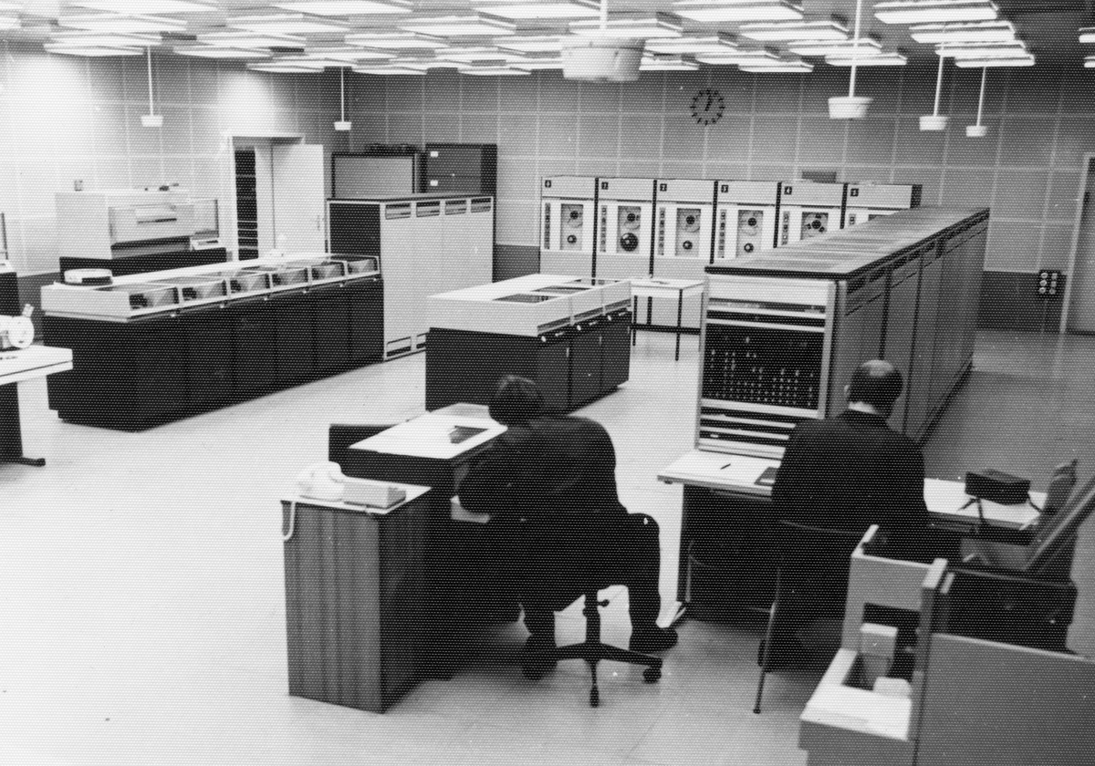 Auf dem ein schwarz-weißen Lichtbild sind zwei Männer am Schreibtisch sitzend und von hinten vor Siemens-Großrechnern S4004/45 im Zentralen Rechenzentrum zu sehen.