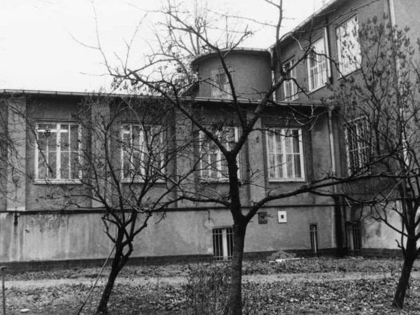 Hinter drei kleinen kahlen Laubbäumen ist die Fensterfront des Anbaus nebst Übergang ins Haupthaus zu sehen. Es handelt sich um ein schwarz-weißes Lichtbild.