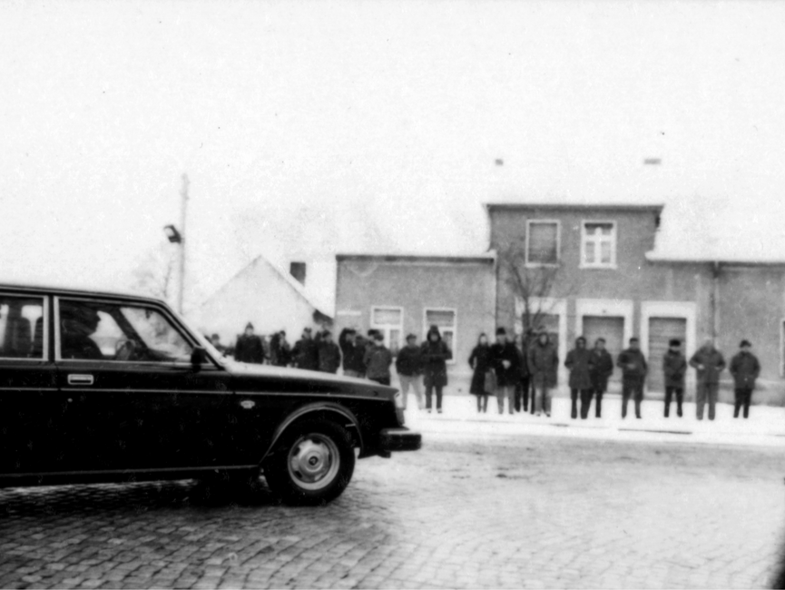 Auf der Schwarzweißaufnahme sind nur einige wenige Zuschauer an der Protokollstrecke in Güstrow während Helmut Schmidts Staatsbesuchs in der DDR, zu sehen. Hinter den Zuschauern ist ein kleines Wohnhaus. Auch hier liegt Schnee. Am linken Bildrand ist ein Auto, das nach rechts einbiegt zu erkennen.
