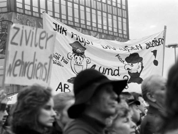 Detailaufnahme einiger Personen, die an einem Demonstrationszug am Berliner Alexanderplatz teilnehmen. Im Vordergrund ein Mann mit Hut und eine Frau, dahinter ein kleines Transparent mit der Aufschrift "ziviler Wehrdienst". Dahinter befindet sich ein größeres Plakat mit der Aufschrift "Ihr müßt draußen bleiben" und Zeichnungen, die auf einen Volkspolizisten und einen Stasi-Mitarbeiter anspielen.