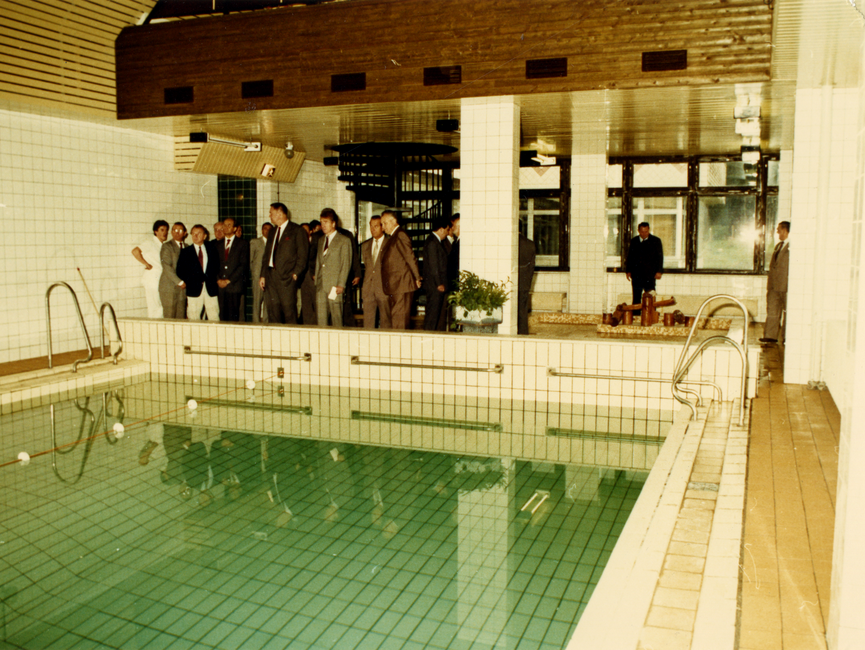 Mehrere Herren im Anzug stehen vor einem Pool und sehen sich um. Es handelt sich um ein farbiges Lichtbild.