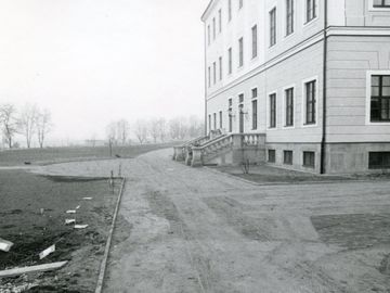 Die Schwarz-Weiß-Fotografie zeigt die Rückseite des Hotels Bellevue in Dresden mit einer zu einem rückwärtigem Eingang führenden Treppe. Das Haus rechts im Bild hat drei sichtbare Stockwerke und wurde mit hellem Sandstein erbaut. Links davon, ausgehend vom Vordergrund und sich beidseitig um das Gebäude ziehend, findet sich ein breiter Sandweg. Gegenüber der Treppe, links im Bild, führt der Weg ab in ein spärlich von Bäumen bewachsenes Brachland.