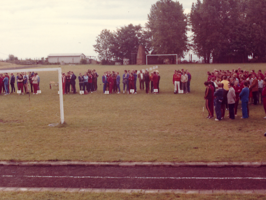 [Das farbige Lichtbild zeigt einen Fußballplatz vor einer Baumgruppe. Gruppenweise sind die Sportler hinter, in den Rasen gerammten, Schildern mit Nummern versammelt.Im linken Bildhintergrund steht eine weiße, eingezäunte Baracke.]