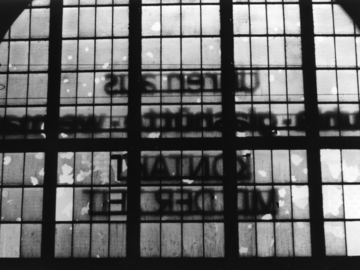Das schwarz-weiße Lichtbild zeigt die Innensicht auf die große Fensterfläche der Bahnhofsfront des Dresdner Hauptbahnhofs. Der Schriftzug ist seitenverkehrt. Viele der kleinen Fenster sind zerstört.
